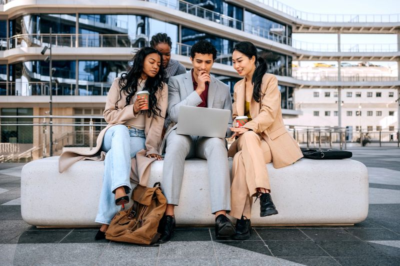 Four business professionals collaborate outdoors in the city while using a laptop. Captures teamwork, mobile work, and a modern urban business lifestyle.