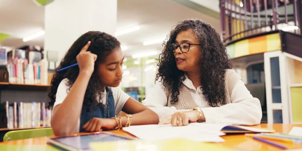 A woman helps a young girl with homework at a library table.