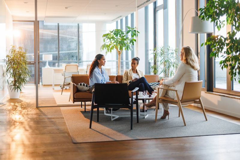 Diverse group of businesswomen having a productive meeting in modern office setting with large windows and natural light, sitting on comfortable chairs around a table, expressing engagement and collaboration.