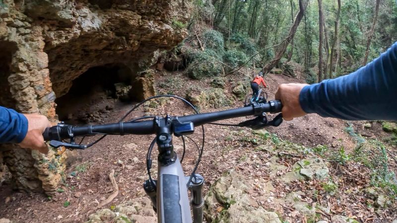 POV from father's hands on handlebars, following adult daughter on electric mountain bike through forest and old rock ruins, Liguria