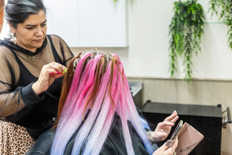 Hairdresser working on a client's hair, applying vibrant pink, blue, and brown synthetic extensions with glitter accents for a new stylish look at a salon