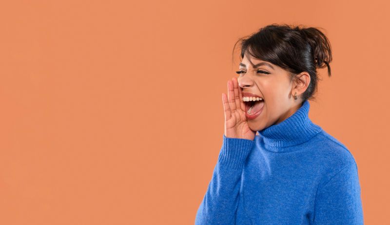 A woman enthusiastically shouts while smiling, showcasing joy against a vibrant orange background, dressed warmly in a stylish blue sweater.
