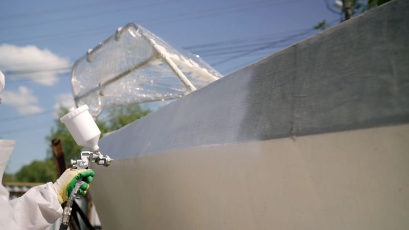 Skilled maritime professional applying protective coating on boat hull using spray gun in shipyard setting, wearing specialized safety equipment during precise maintenance process