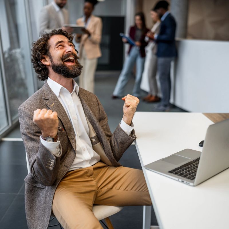 Mature adult man with a beard cheering excitedly, looking up with fists clenched in a gesture of triumph in an office
