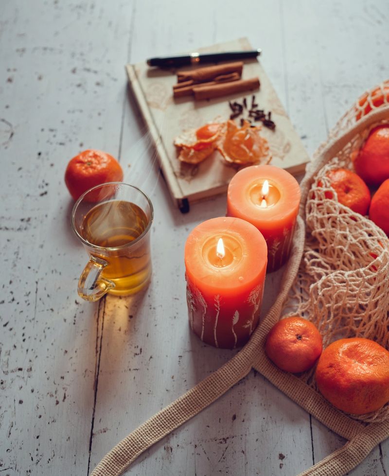 Cozy winter still life featuring a glass of hot tea, glowing candles, fresh tangerines in a reusable mesh bag, and a notebook with spices on a white wooden background. Warm light, natural textures, and intentional styling create a calm seasonal atmosphere with generous copy space.