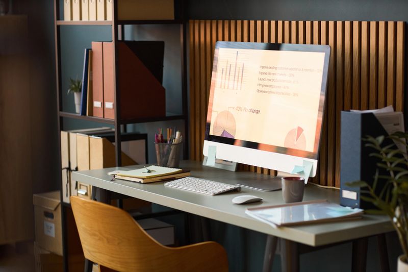 Modern workspace featuring desktop computer displaying business analytics charts and graphs on screen, organized desk with office supplies, shelving unit with folders and documents in background