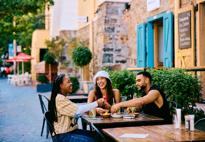 Group of joyful friends sitting at an outdoor cafe, smiling and raising drinks while enjoying food and conversation during a relaxed city travel experience.