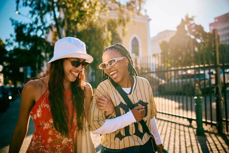 Two stylish young women walking side by side on a city street, smiling and laughing together, sharing a carefree and friendly moment during an urban outing.