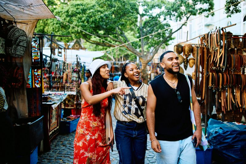 Group of cheerful young tourists walking through a city street market, smiling and talking as they browse stalls and enjoy a social travel experience.