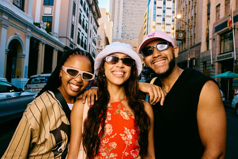 Portrait of three stylish friends standing together on a city street, smiling at the camera and sharing a friendly and relaxed travel experience outdoors.