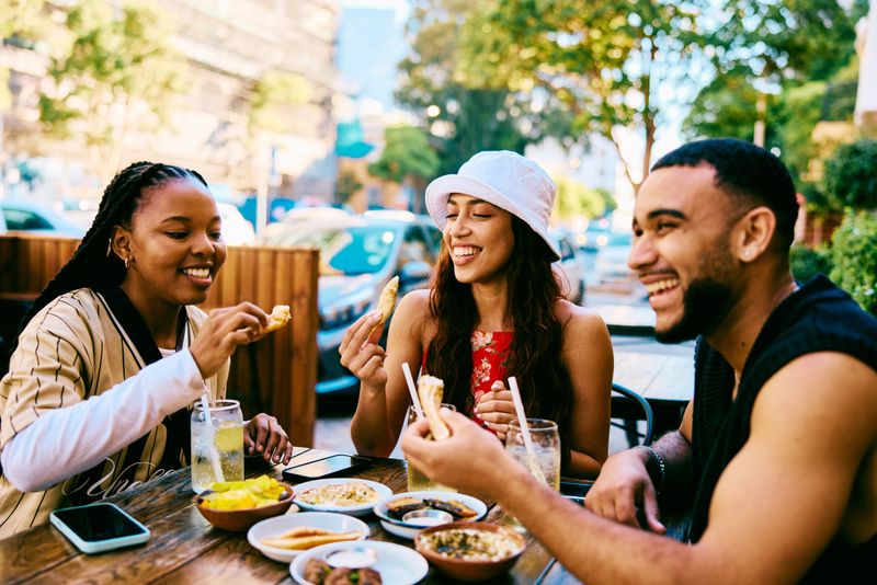 Candid shot of happy young adults sitting around a cafe table outdoors, laughing and sharing food while enjoying relaxed conversation in a vibrant urban street.