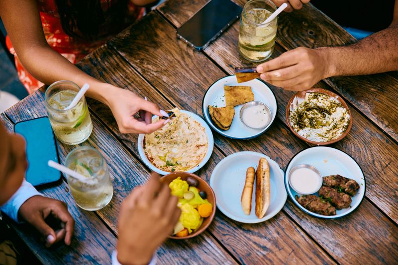 Overhead view of people's hands reaching for shared plates of vibrant food and drinks on a restaurant table, showing connection, variety, and social dining.