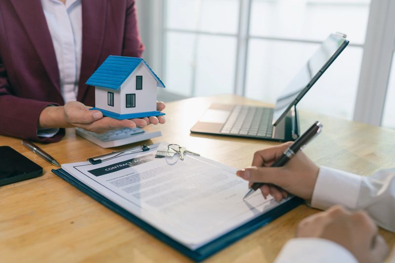 A real estate agent or broker is showing a house plan to a client. The plan includes a pen, and they are discussing contract details with the client during a meeting in the office.