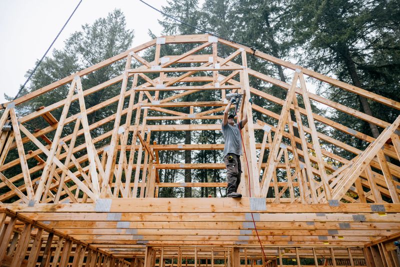 Workers build the frame of a new house project. Bare plywood and beams as it is framed up from the foundation. High lumber costs have affected the building process.  A crane lowers assembled rafters and trusses.  Shot in Washington state, USA.