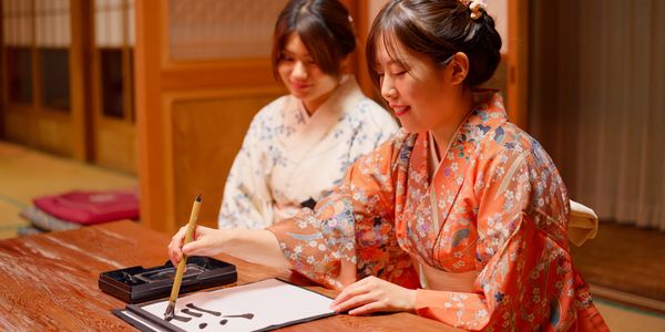 Two women in kimonos practicing Japanese calligraphy indoors.
