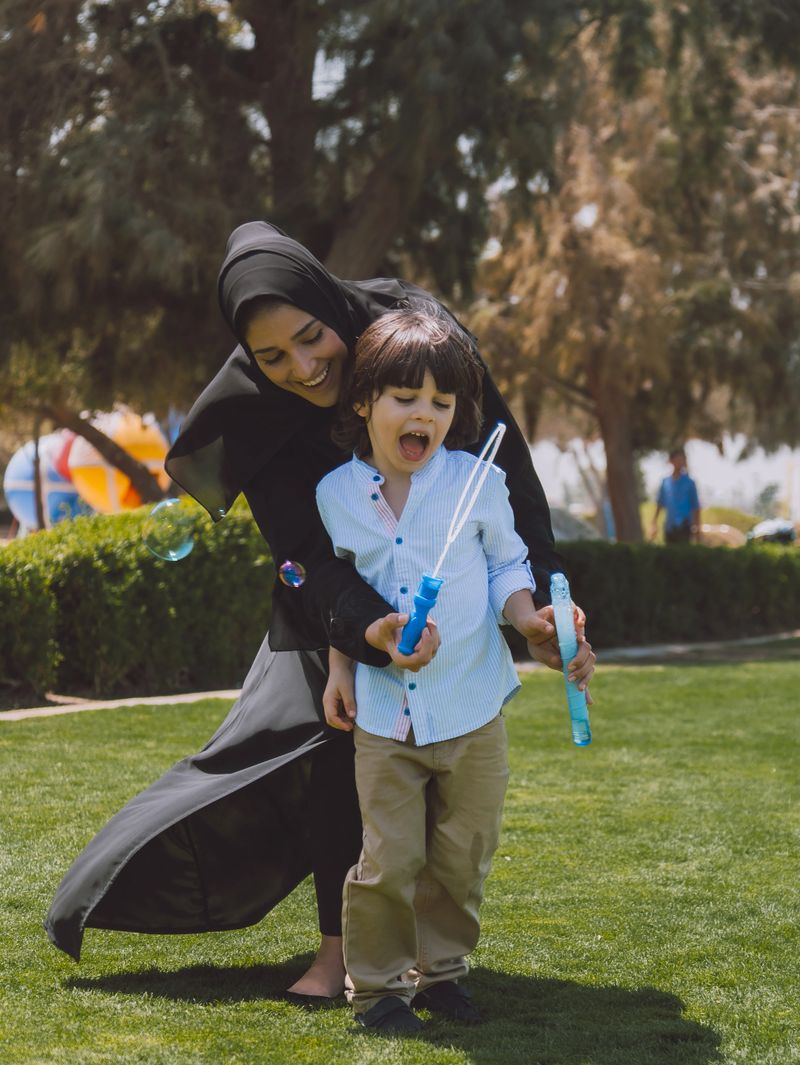 Happy Arab mother wearing a black abaya plays soap bubble game with her young son in a sunny public park in Dubai, United Arab Emirates. The cheerful child enjoys blowing and chasing bubbles while spending quality family time outdoors during a warm day, capturing joyful parent-child interaction, childhood happiness, and relaxed Middle Eastern family lifestyle