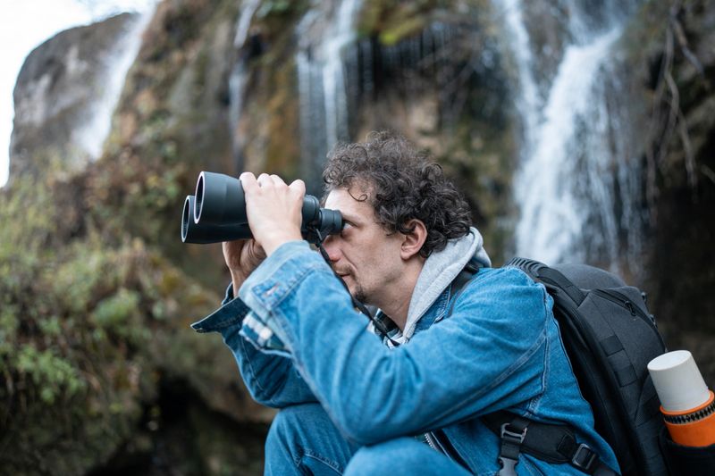 A curious photographer crouches beside a waterfall, using binoculars to scout the terrain. He wears a denim jacket and backpack, ready for outdoor exploration and nature photography in a rugged landscape.