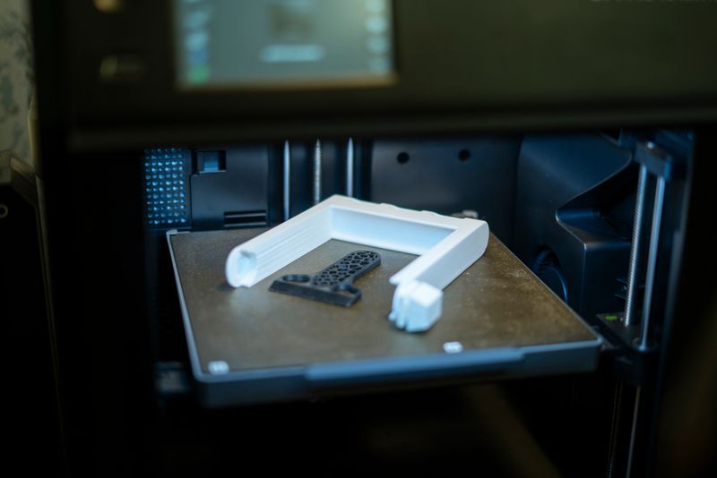 A 3D printer is in action, producing a white item with intricate designs on a build plate in a well-lit workshop, showcasing advanced printing technology.