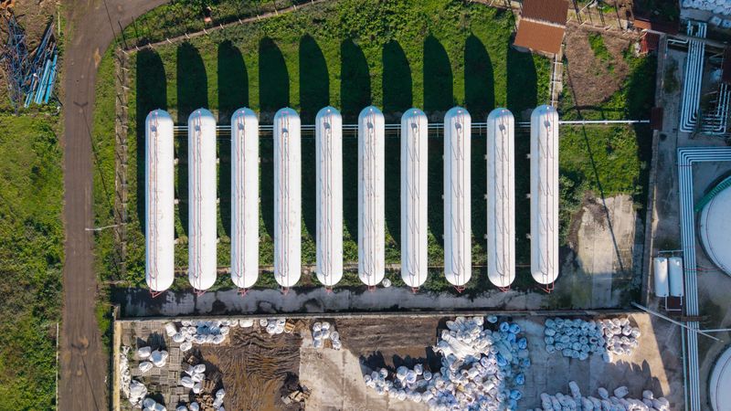 An overhead drone shot captures a series of large white cylindrical storage tanks arranged neatly in an industrial facility with green grass and shadows