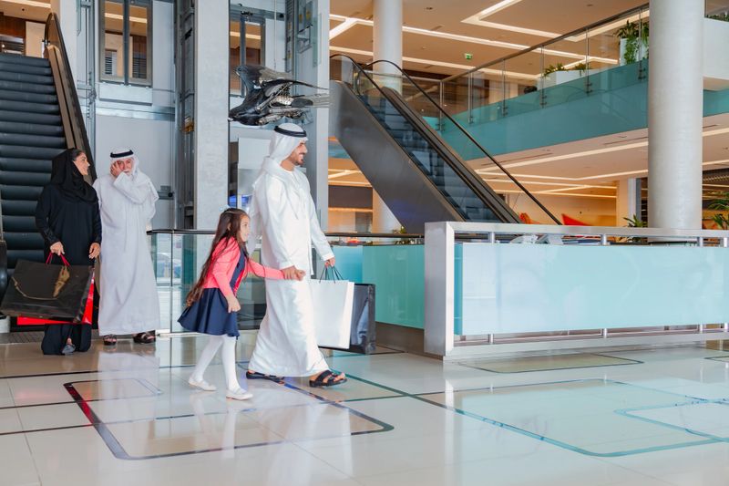 A Middle Eastern three generation family consisting of an elderly grandfather, adult parents, and a young girl walk together inside a modern shopping mall while holding hands, expressing family bonding, care, and togetherness. The grandfather wears traditional white kandura and headscarf while talking on a smartphone, the mother wears a black abaya and carries shopping bags, and the little girl wears a navy dress with a pink cardigan. The scene is captured in a bright contemporary mall corridor with glass railings, indoor plants, polished floor reflections, and a cafe and patisserie in the background, representing modern lifestyle, family values, retail experience, and everyday life in the Middle East
