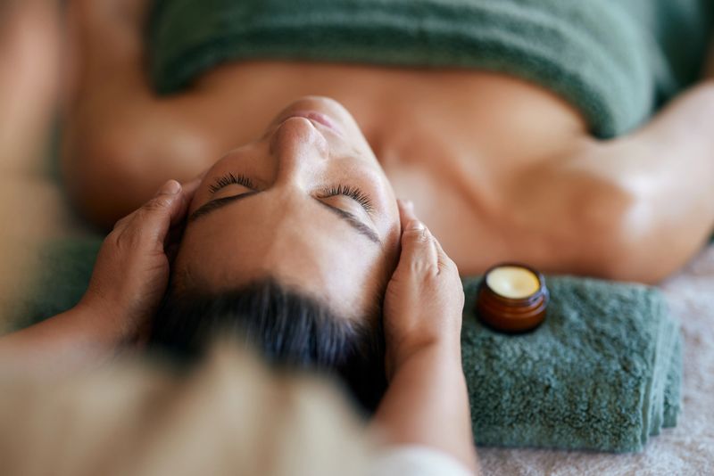 Therapist massaging woman face with both hands using soothing cream. Close up face of latin woman lying calmly getting facial treatment at luxury spa with closed eyes. Beautiful hispanic girl pampering self at massage therapy center.