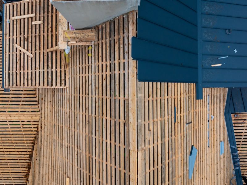 Aerial view shows a pitched roof with wooden battens and dark metal cladding, loose timber offcuts, and workers on a residential site in daylight during mid stage work.
