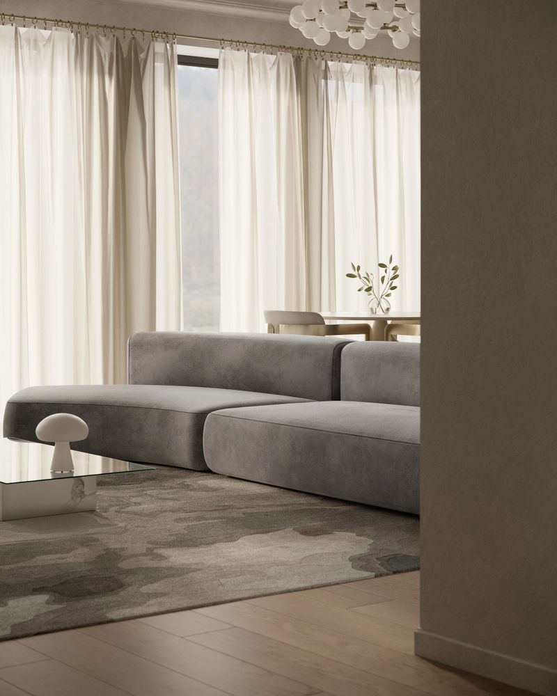 A curved gray modular sofa sits on a textured designer rug, complemented by a minimalist glass coffee table and sculptural decorative elements. The open-plan kitchen features muted blue cabinets, marble countertops, and integrated appliances. Decorative ceiling moldings and a modern chandelier lend the space a sophisticated, timeless character.