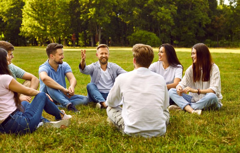 Friendship and communication. Group of young people communicate sitting on grass in park on summer sunny day. Group of male and female friends laughing and talking while sitting in circle outdoors.