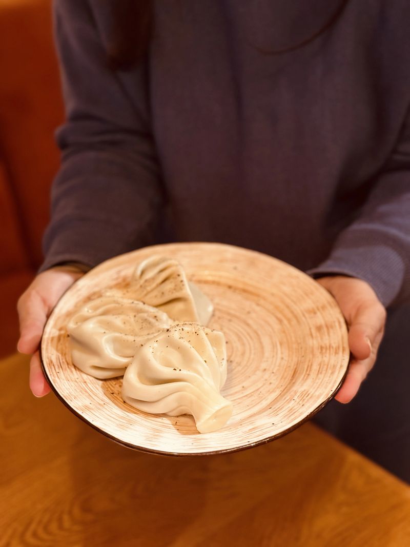 Hands Holding Plate With Dumplings, Warm Lighting And Cozy Restaurant Vibe, Ceramic Dish With Three Pleated Steamed Dumplings Offered By Server, Rustic Wood Table Background And Textured