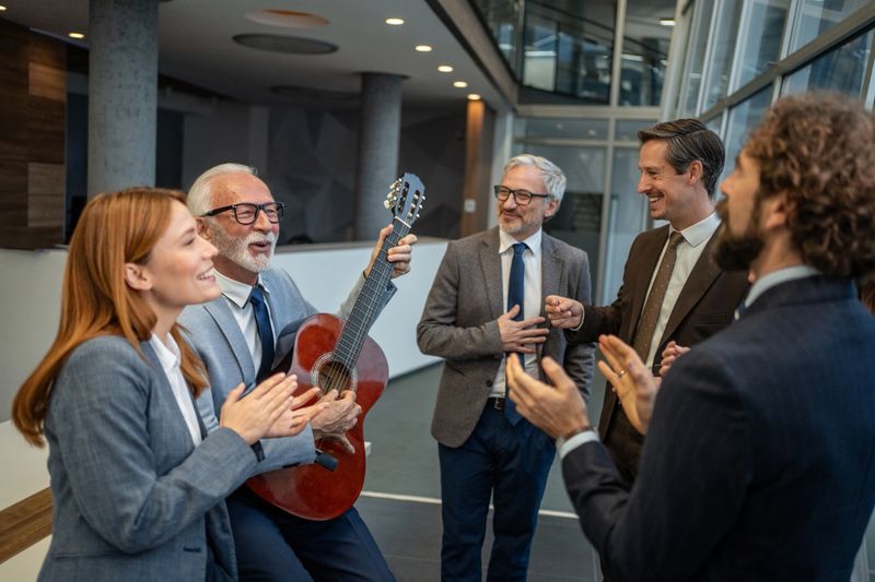 Diverse business professionals celebrating a joyful office party. A senior adult playing guitar while colleagues smile. Clap. And enjoy the lively musical performance. Fostering teamwork and happiness