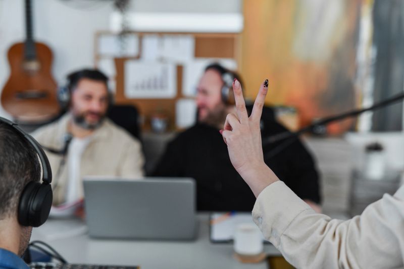 A lively podcast recording session with hosts and guests wearing headphones around a desk, laptop, and microphones in a bright studio. A guest makes a peace sign while the conversation flows.