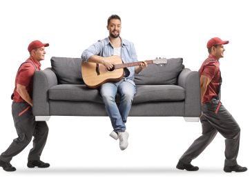 Man playing guitar on a sofa carried by two movers.