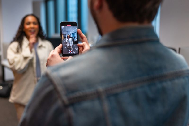 Young Hispanic woman in modern office energetically performing for smartphone camera held by colleague, creating engaging content with joyful expression and enthusiasm.