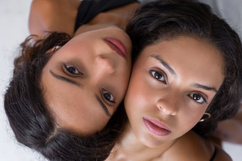 Close up portrait of twin sisters lying together and looking at the camera. Natural light, emotional bond, sisterhood, beauty and intimacy concept for lifestyle, wellness and advertising use.