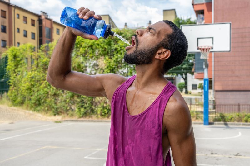 Mid adult multiracial man enjoying a refreshing drink from a water bottle on a street basketball court, wearing a purple tank top. The outdoor setting features urban buildings and a basketball hoop in the background, highlighting a relaxed and joyful moment.
