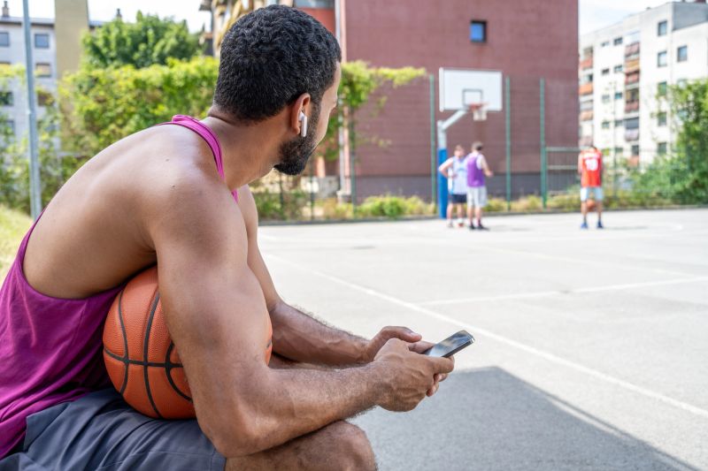 Mid adult multiracial man sitting on an urban basketball court, holding a basketball and using a smartphone while waiting for his turn to play under the bright afternoon sun.