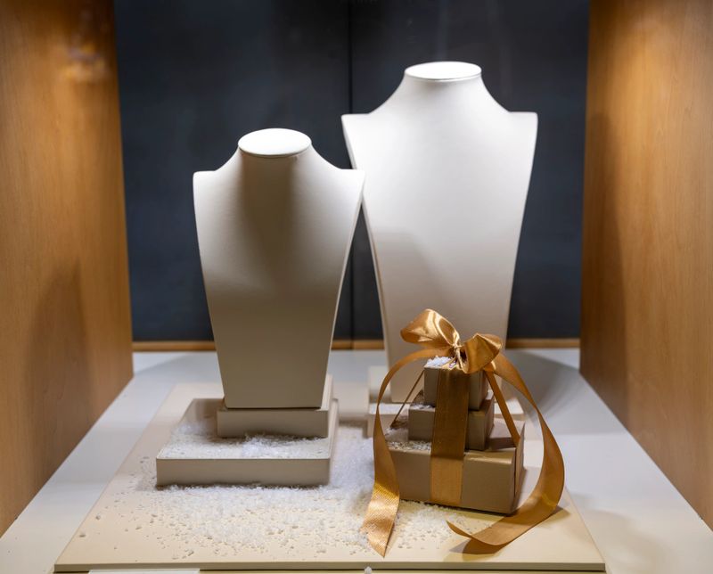 Two white jewelry display busts stand side by side on a white surface with textured details beside a small gift box wrapped in gold ribbon in a softly lit setting