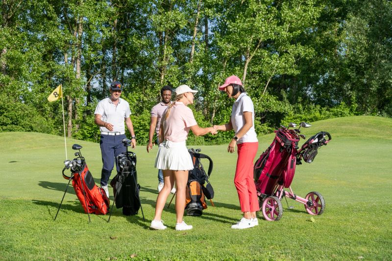 Diverse male and female golfers enjoying a lively interaction near their golf trolleys on a lush course, with bright sunlight enhancing the outdoor setting. The group appears cheerful and engaged, embodying the spirit of friendly competition.