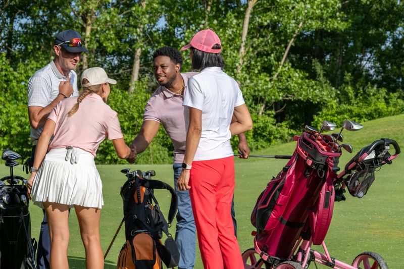 Diverse male and female golfers gathered on a sunlit course, engaging in friendly conversation next to their golf trolleys. The outdoor setting is vibrant with lush greenery surrounding them, as they enjoy the camaraderie of the game.