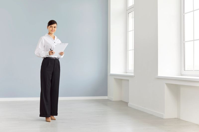 Portrait of confident smiling young woman real estate agent agent standing in new empty apartments with documents in hands and looking cheerful at camera. Real estate market and moving day concept.