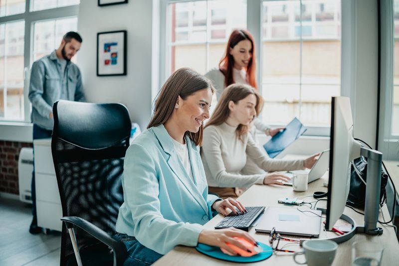 Three coworkers collaborate on tasks at their desks in an office. Two women focus on their computers while a man works in the background. There are documents and coffee cups on the table.