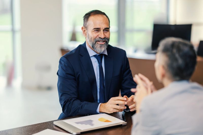 Business professional in a suit consulting with a client at an office table, discussing financial planning, documents and strategy during a serious business meeting