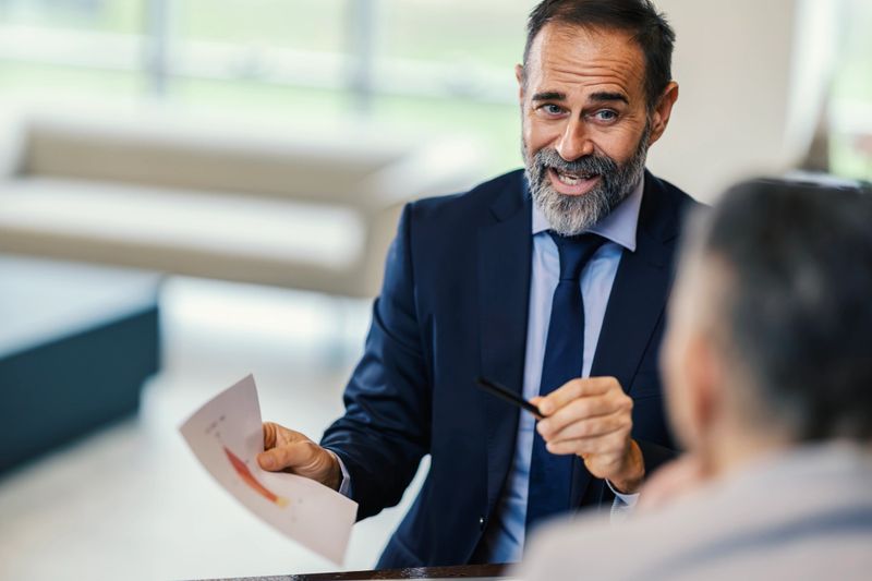 Mature adult businessman wearing a suit and tie confidently discussing financial charts and growth data with a client during a successful professional meeting in a modern office environment
