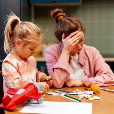 Child playing with a craft at a table. Mom with her hand on her forehead looking down. Mom is sittin