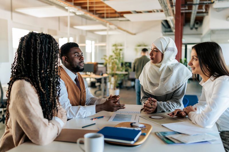 Colleagues sit around a table in a modern office, actively discussing ideas during a team meeting. The scene highlights collaboration, communication, and teamwork in a contemporary workplace.