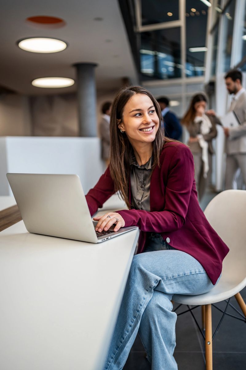 Young adult woman smiling and typing on a laptop, enjoying a productive day in a contemporary business office