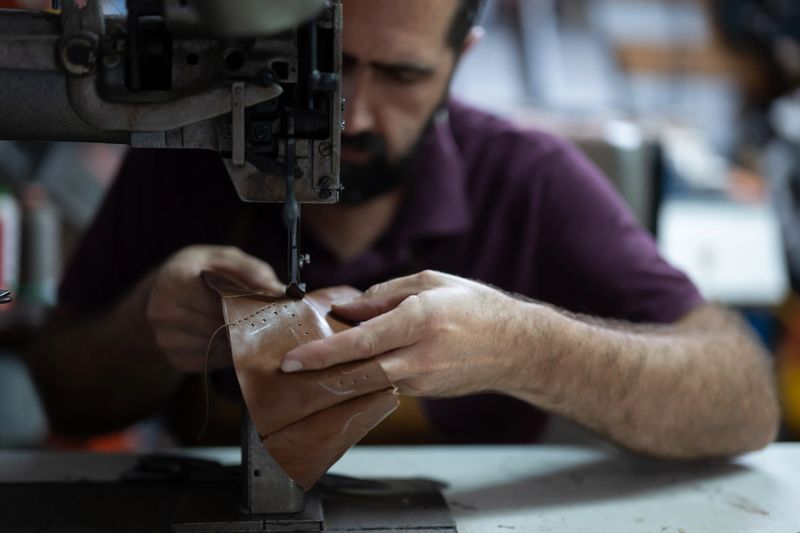 A craftsman concentrates on stitching a leather item at an industrial sewing machine in a busy workshop. Close-up of hands and thread highlights skill, precision, and handmade quality.