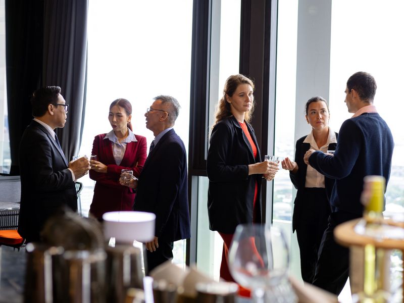 Business professionals networking at corporate event holding drinks and discussing ideas near large windows;collaboration communication confidence and professional social interaction