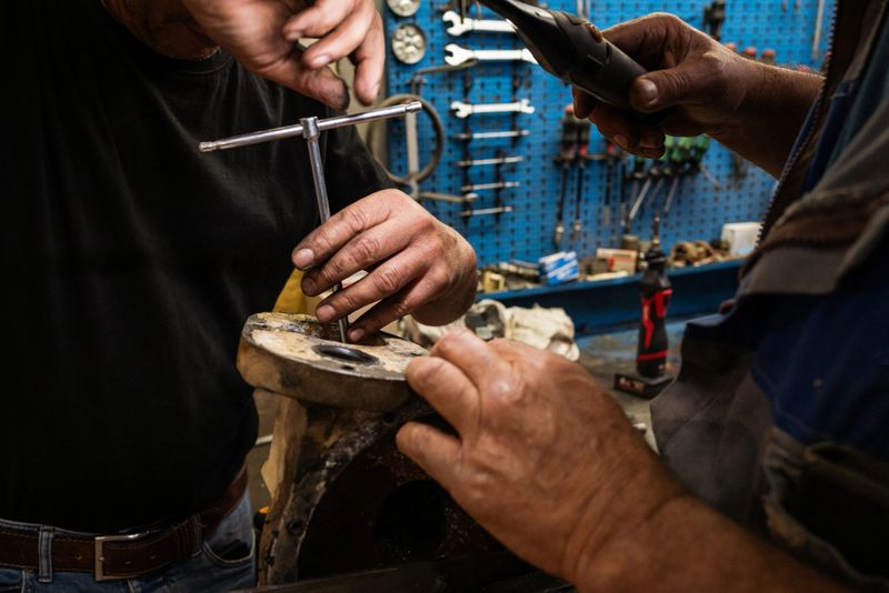 Close-up of a mechanic’s hands using a caliper and hand tools to measure and repair a metal mechanical component inside a workshop. The image highlights precision, craftsmanship, and manual labor in an industrial maintenance environment, with tools and equipment visible in the background.