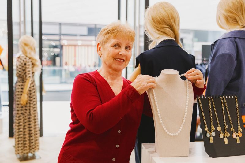 Portrait of one beautiful happy elderly caucasian woman in red blouse taking pearl necklace hanging on stand to buy in shop on spring day, side view close up. Concept of shopping time, retirement lifestyle, choosing jewelry.
